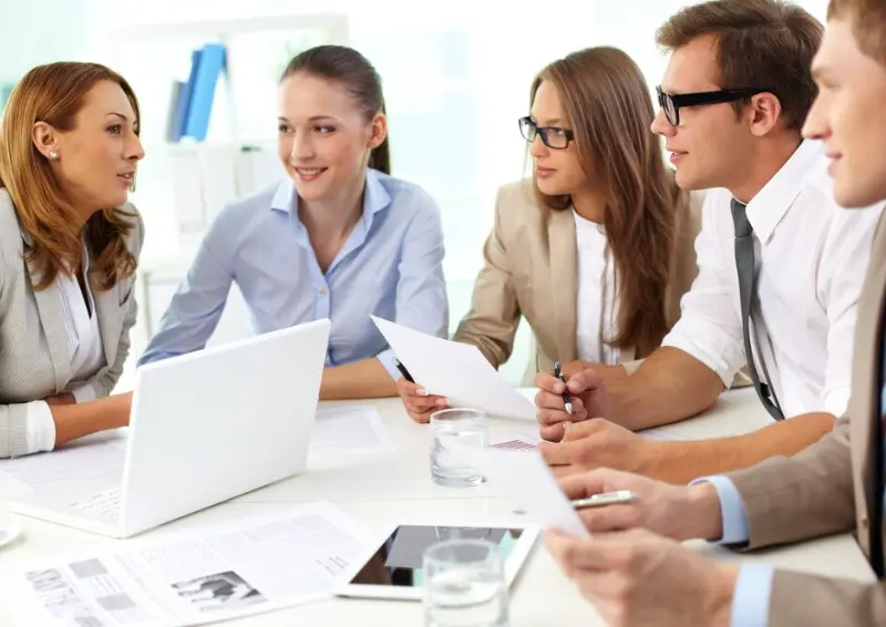 A group of business professionals seated around a table, reviewing documents and a laptop